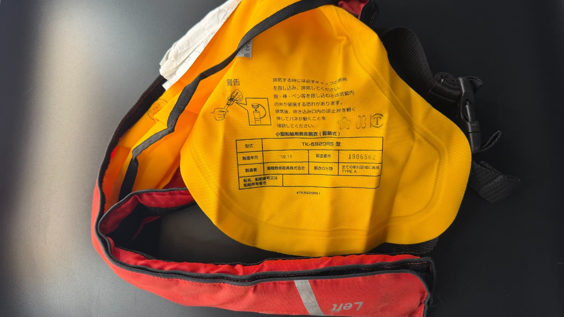 膨張式ライフジャケットの気室に印字された型式承認ラベル。桜マークと JG マーク、型式 TK-5920RS、製造者 高階救命器具株式会社、胴衣の分類「全ての航行区域に適用 TYPE A」の表